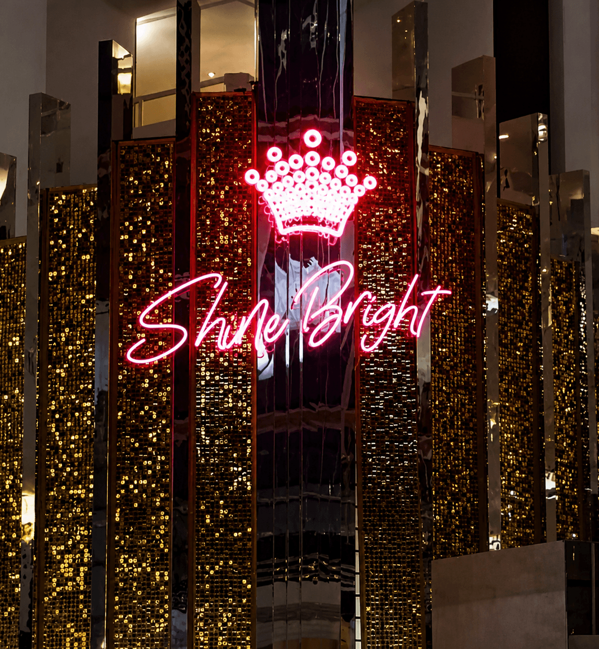 Pink script neon sign reading Shine Bright under a crown, on a sequin wall