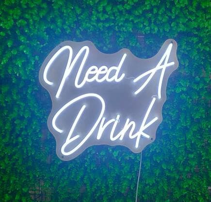 White script neon sign reading Need A Drink on a greenery wall